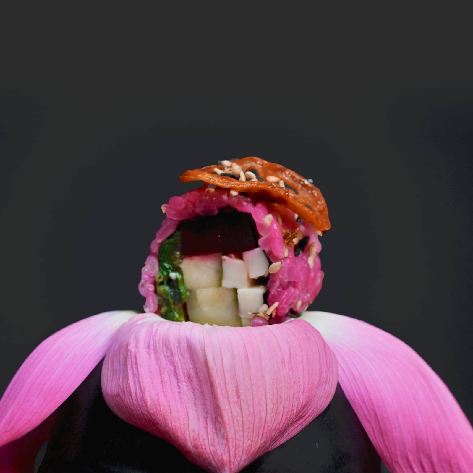 Pink rice sushi piece with cubed filling and a sesame crisp, resting on lotus petals